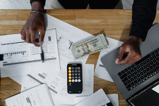 Hands calculate finances with papers, cash, and a laptop on a wooden desk.